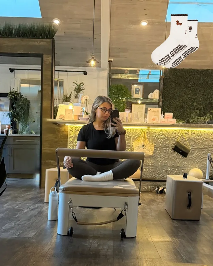 A mirror selfie cross-legged on a reformer in Weenie and Espresso Martini grip socks